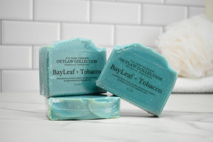 Three bars of soap labeled 'BayLeaf + Tobacco' from the Outlaw Collection on a white surface with a tiled wall background.