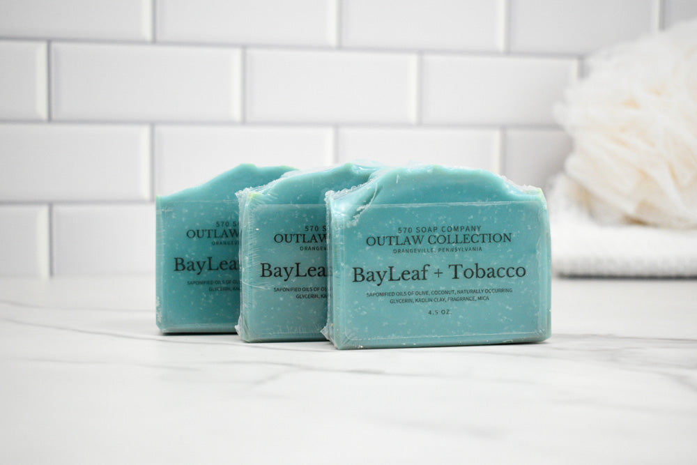 Three bars of soap labeled 'BayLeaf + Tobacco' from the 'Outlaw Collection' on a white surface with a tiled wall background.