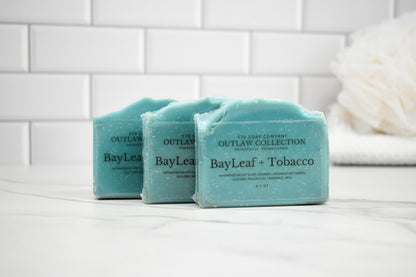 Three bars of soap labeled 'BayLeaf + Tobacco' from the 'Outlaw Collection' on a white surface with a tiled wall background.