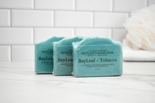 Three bars of soap labeled 'BayLeaf + Tobacco' from the 'Outlaw Collection' on a white surface with a tiled wall background.