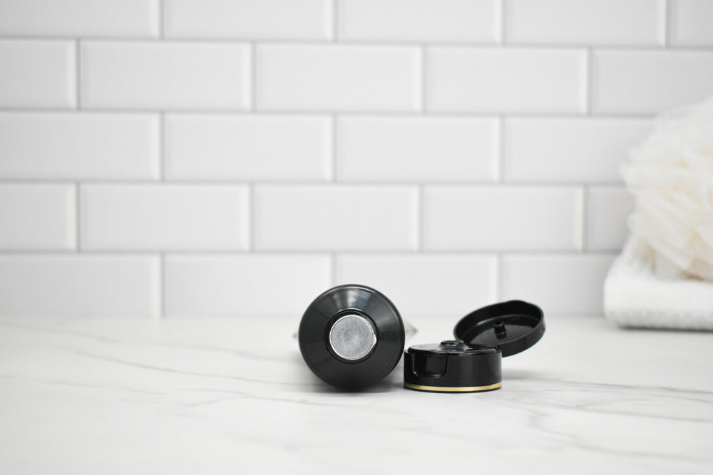 Black and gold lotion bottle and lid on a white surface with a tiled wall background