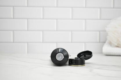 Black and gold lotion bottle and lid on a white surface with a tiled wall background