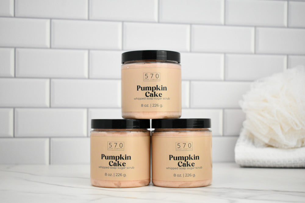 Three jars of 570 Pumpkin Cake body scrub on a white surface with a tiled wall background.
