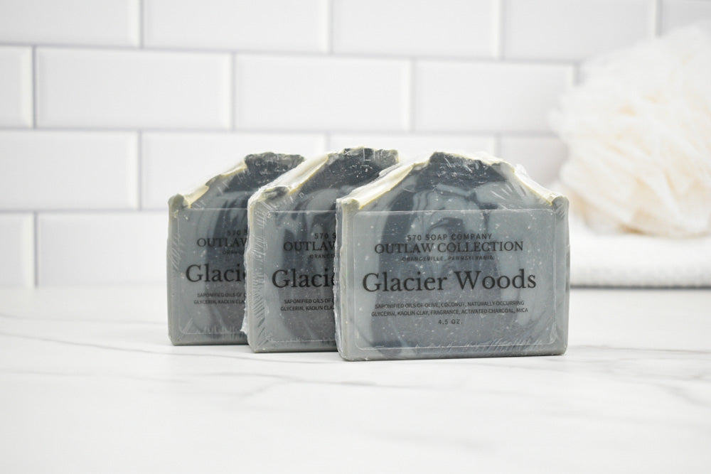 Three bars of 'Glacier Woods' soap from The Soap Den on a white surface with a tiled wall background.