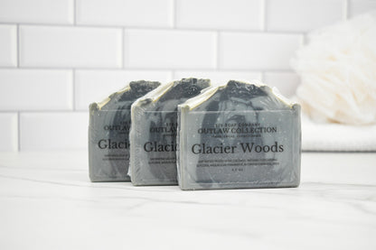 Three bars of 'Glacier Woods' soap from The Soap Den on a white surface with a tiled wall background.