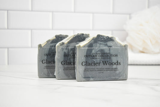 Three bars of 'Glacier Woods' soap from The Soap Den on a white surface with a tiled wall background.