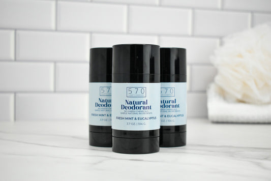 Three 570 Natural Deodorant sticks on a white surface with a tiled wall background