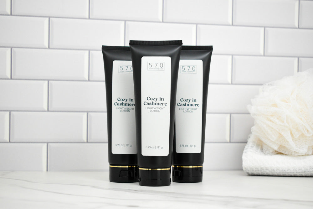Three black and white tubes of Cozy in Cashmere lotion on a white surface with a tiled wall background.