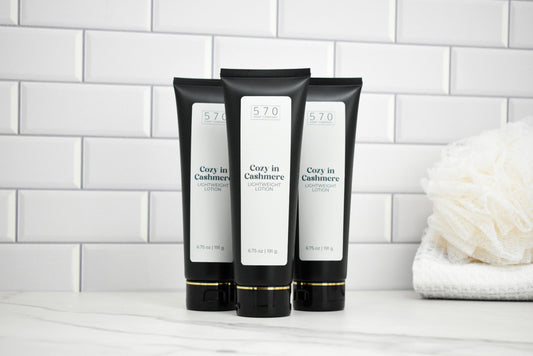 Three black and white tubes of Cozy in Cashmere lotion on a white surface with a tiled wall background.