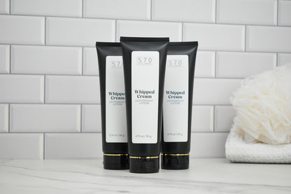 Three black tubes of Whipped Cream lotion on a white surface with a tiled wall background.