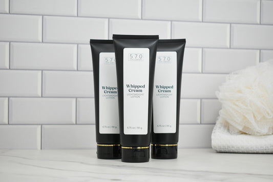 Three black tubes of Whipped Cream lotion on a white surface with a tiled wall background.