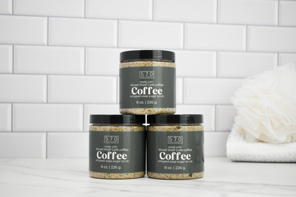 Three jars of coffee scrub stacked on a white surface with a tiled wall background