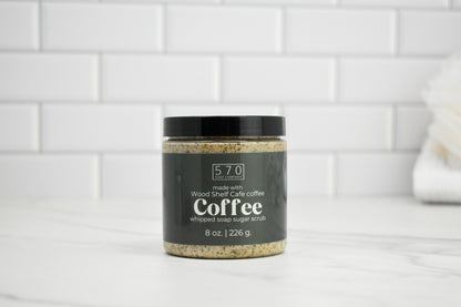 Jar of coffee-flavored whipped soap sugar scrub on a white surface with a tiled wall background