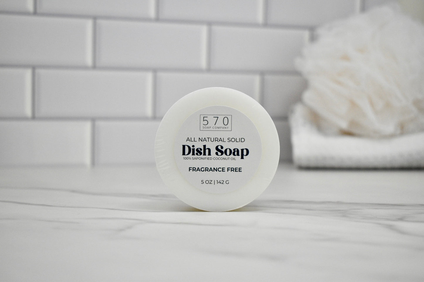 All natural solid dish soap container on a bathroom counter with white tiles in the background.
