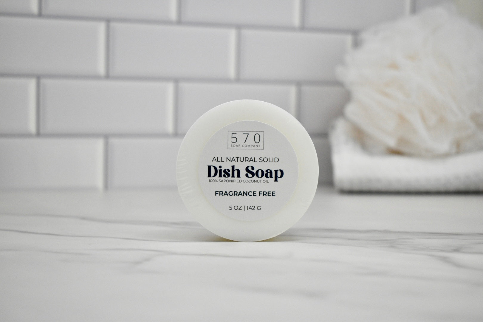 All natural solid dish soap container on a bathroom counter with white tiles in the background.