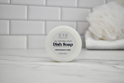 All natural solid dish soap container on a bathroom counter with white tiles in the background.