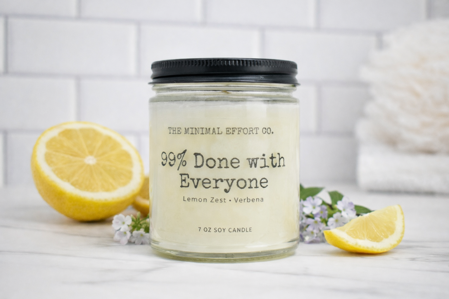 Candle jar labeled 'The Minimal Effort Co.' with lemons and flowers on a white surface.