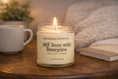 Candle labeled '99% Done with Everyone' from 'The Minimal Effort Co.' on a wooden surface with a cup and book in the background.