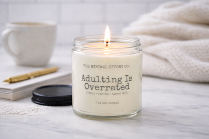 Candle with 'The Minimal Effort Co.' branding on a table with a mug, pen, and notebook.