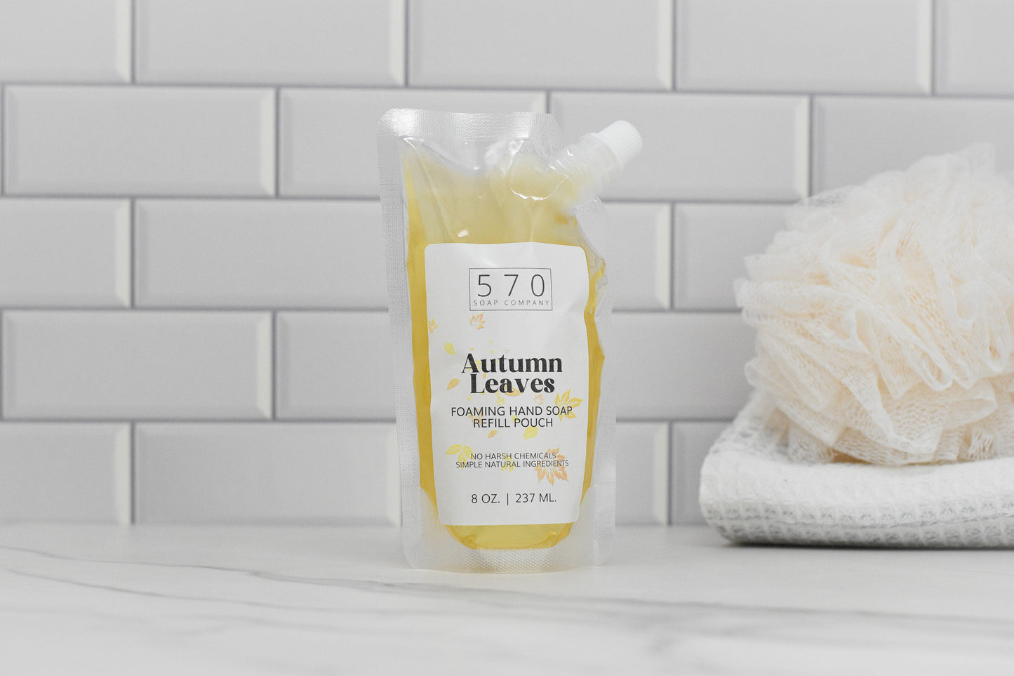 An 8 oz (237 mL) refill pouch of Autumn Leaves Scented Foaming Hand Soap from 570 Soap Co., with its delightful Fall Season Aroma, is placed on a countertop against a white tile backsplash. Nearby, you can see a white shower sponge and folded washcloths.