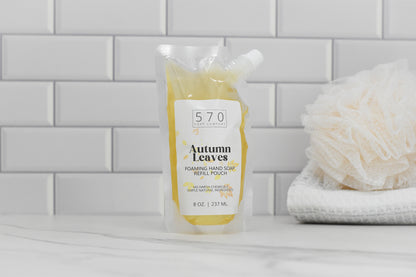 An 8 oz (237 mL) refill pouch of Autumn Leaves Scented Foaming Hand Soap from 570 Soap Co., with its delightful Fall Season Aroma, is placed on a countertop against a white tile backsplash. Nearby, you can see a white shower sponge and folded washcloths.