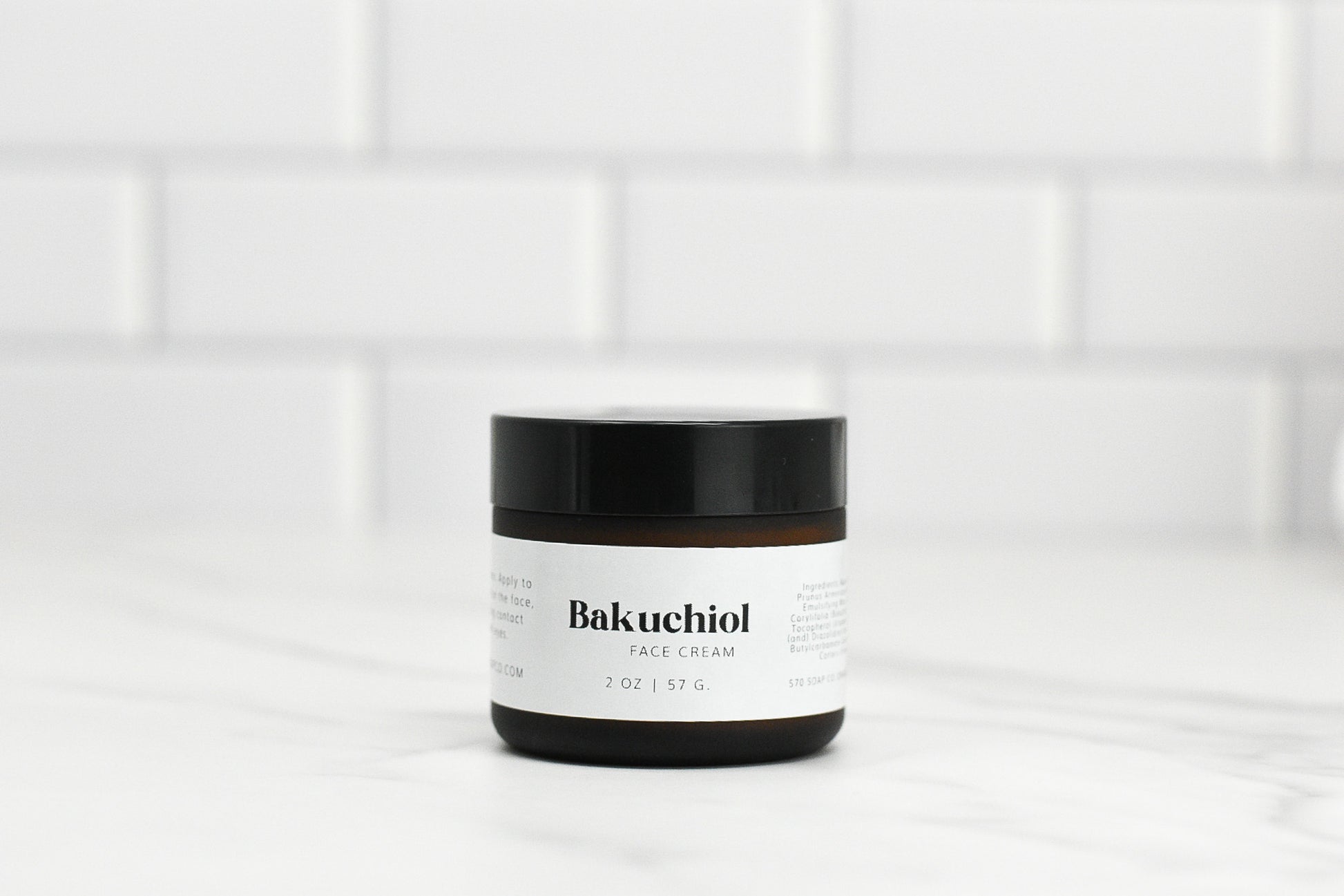 A jar of Bakuchoil Hydrating Face Cream from 570 Soap Co., a must-have in any skincare routine, rests elegantly on a white marble surface. The label stands out against the backdrop of stylish white subway tiles, promising natural radiance with every application.