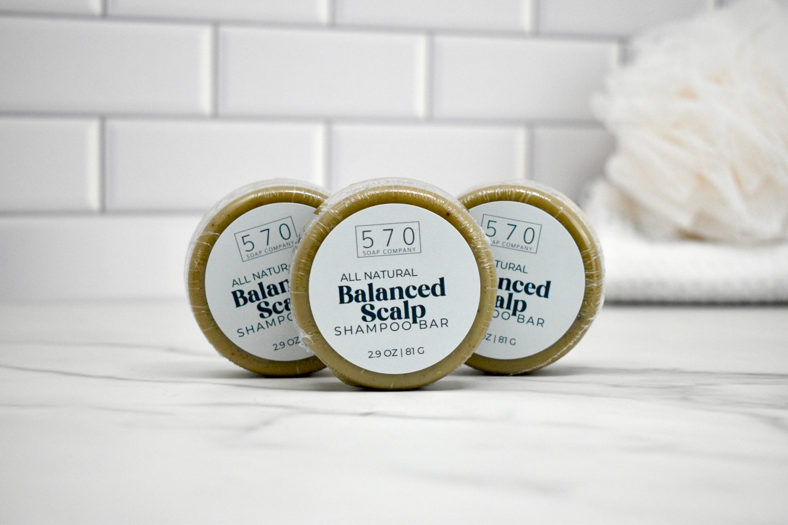 Three round containers of 570 Balanced Scalp Shampoo on a white surface with a tiled wall background.