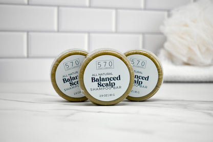 Three round containers of 570 Balanced Scalp Shampoo on a white surface with a tiled wall background.