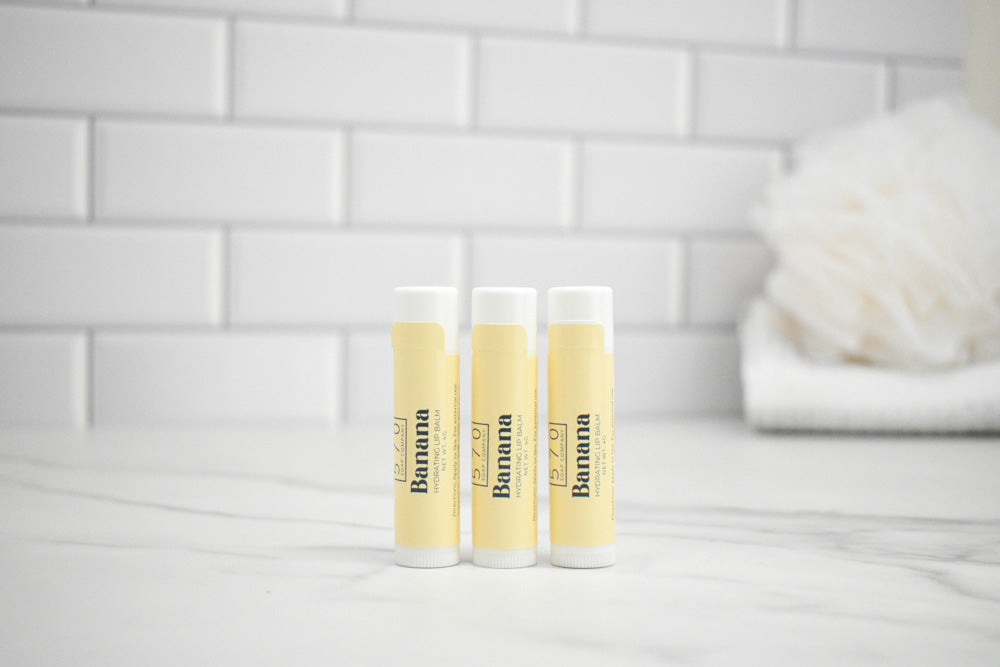 Three lip balm tubes with 'Banana' branding on a white surface with a tiled wall background.