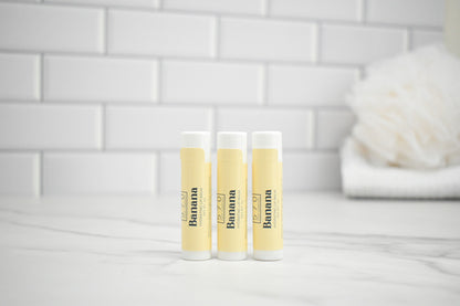 Three lip balm tubes with 'Banana' branding on a white surface with a tiled wall background.