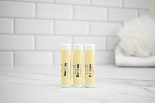 Three lip balm tubes with 'Banana' branding on a white surface with a tiled wall background.