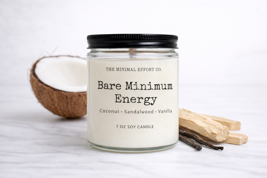 Candle labeled 'Bare Minimum Energy' from The Minimal Effort Co. with a coconut, sandalwood sticks, and vanilla beans on a white background.