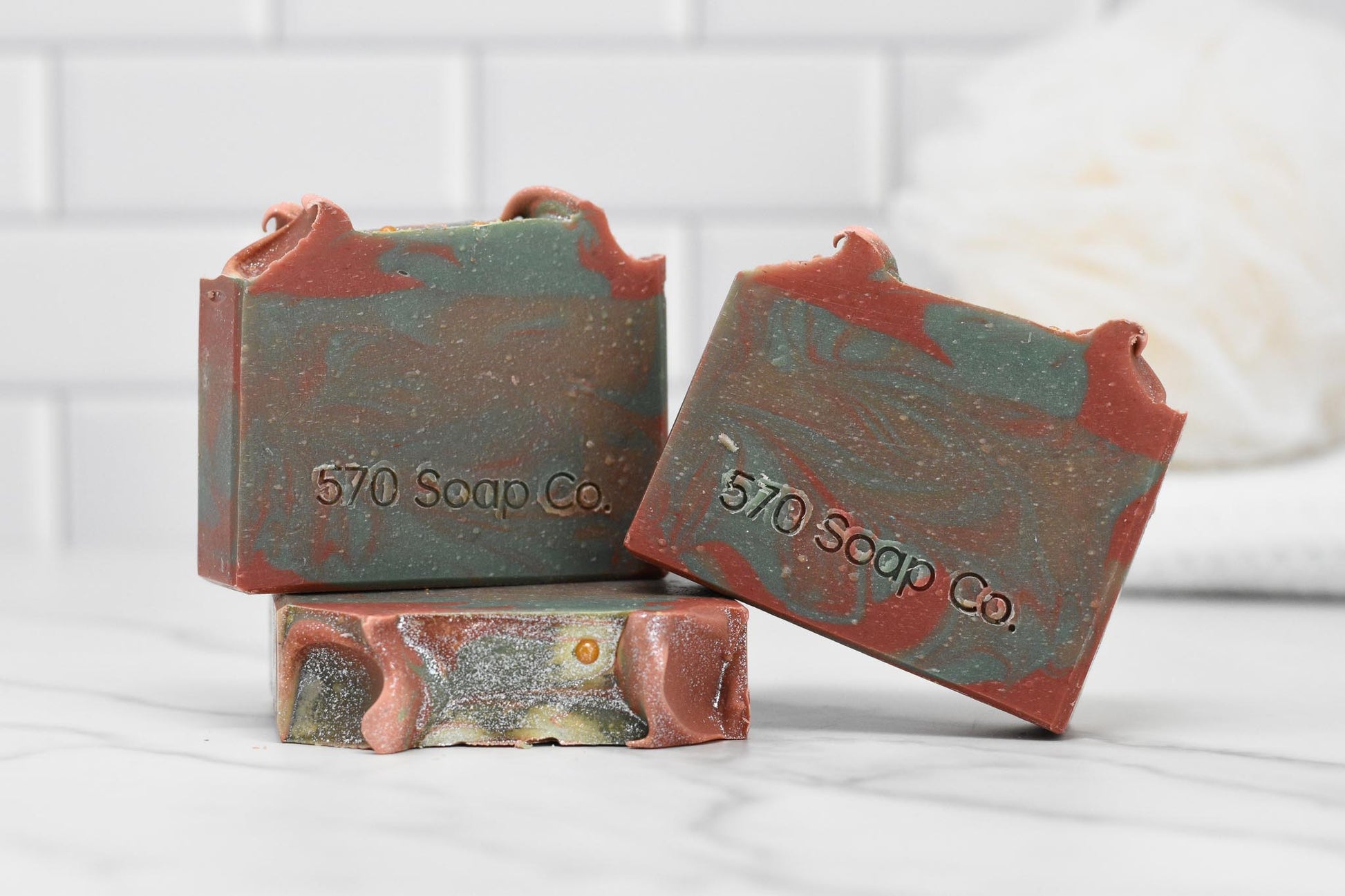 Three Cherry Tobacco Bar Soaps from 570 Soap Co. with a marbled green and brown design are showcased on a white marble surface. Two bars rest against a third, which stands upright. The brand "570 Soap Co." is embossed on them, suggesting the enticing Cherry Tobacco scent. A tiled background enhances the rustic appeal.