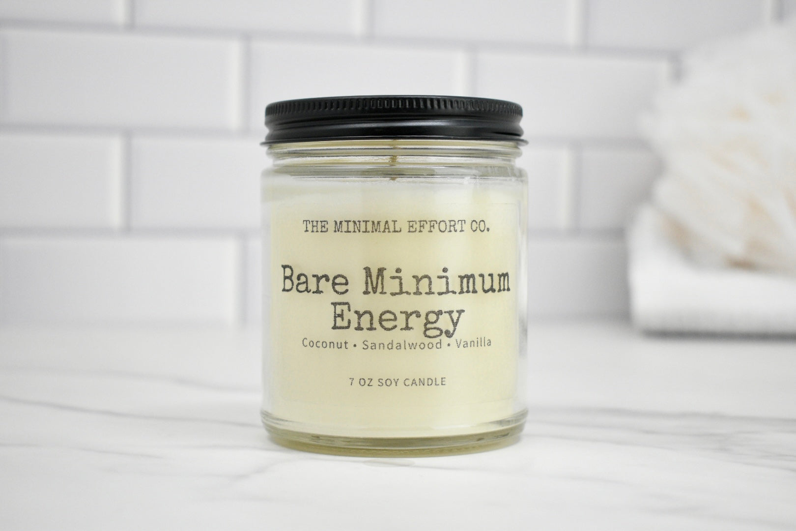 Candle jar labeled 'Bare Minimum Energy' by The Minimal Effort Co. on a white surface with a tiled wall background.