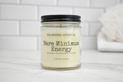 Candle jar labeled 'Bare Minimum Energy' by The Minimal Effort Co. on a white surface with a tiled wall background.