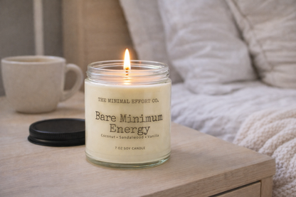 Candle labeled 'The Minimal Effort Co. Bare Minimum Energy' on a wooden surface with a cup and blanket in the background.
