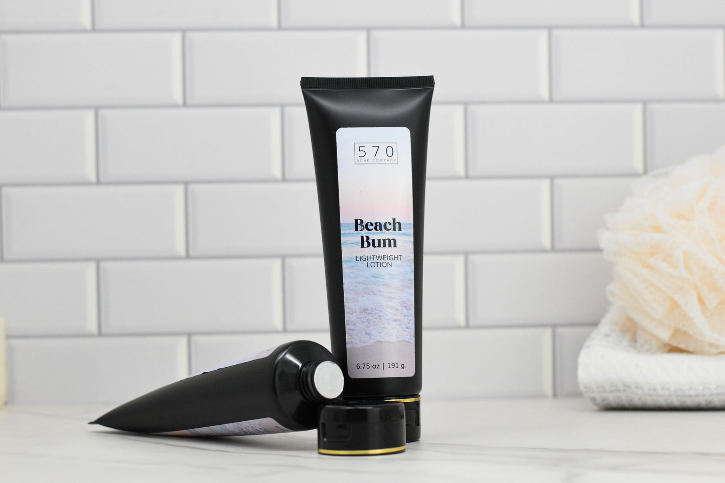 A tube of 570 Soap Co.'s Beach Bum Lightweight Lotion stands upright on a marble surface, promising hydrated skin reminiscent of a tropical paradise. Beside it rests another tube against the backdrop of white subway tiles and a beige bath sponge. Each tube contains 6.75 ounces or 191 grams of lotion.