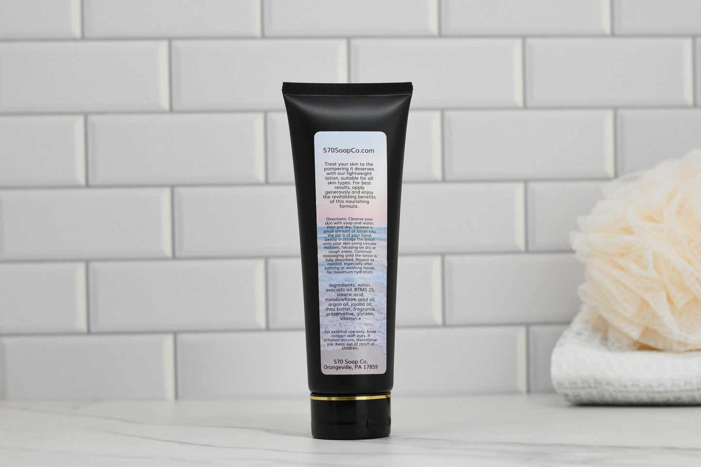 A black tube of 570 Soap Co.'s Beach Bum Lightweight Lotion stands upright on a marble surface against a white subway tile background. A beige loofah and folded white towels evoke a tropical paradise, creating a clean, spa-like ambiance perfect for achieving refreshed and hydrated skin.