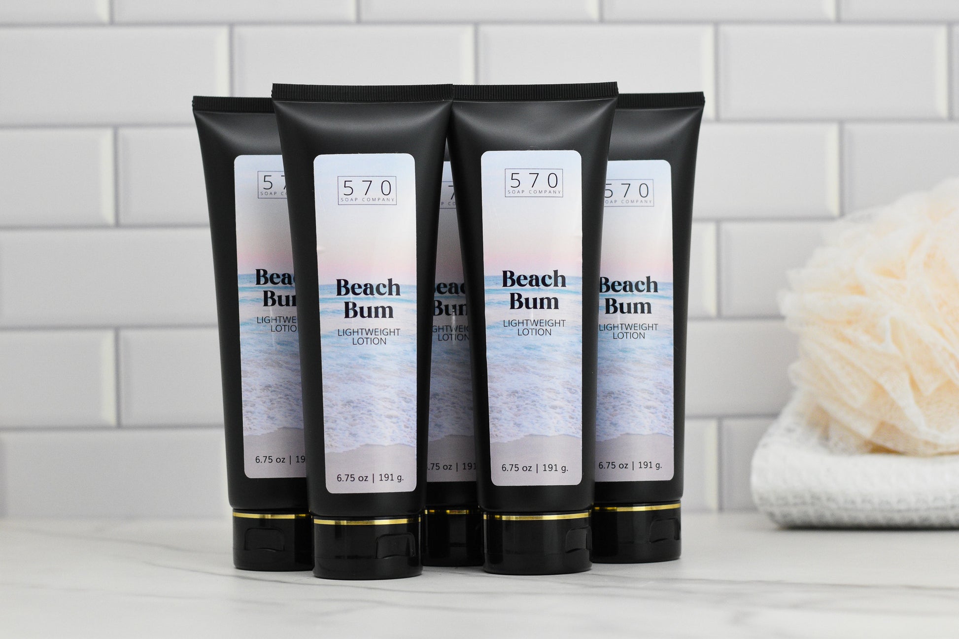 Five tubes of 570 Soap Co.'s Beach Bum Lightweight Lotion are arranged on a marble surface in front of a white tiled wall, capturing the essence of a tropical paradise. Nearby, there's a folded towel and bath sponge. The packaging showcases a coastal image along with a 6.75 oz label, ensuring perfectly hydrated skin.