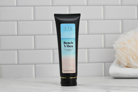 A black tube of 570 Soap Co.'s Beach Vibes Lightweight Lotion, enriched with skin-loving ingredients, stands on a marble surface against a white tile background. Partially visible on the right side is a beige bath sponge that evokes the feeling of a tropical beach breeze.