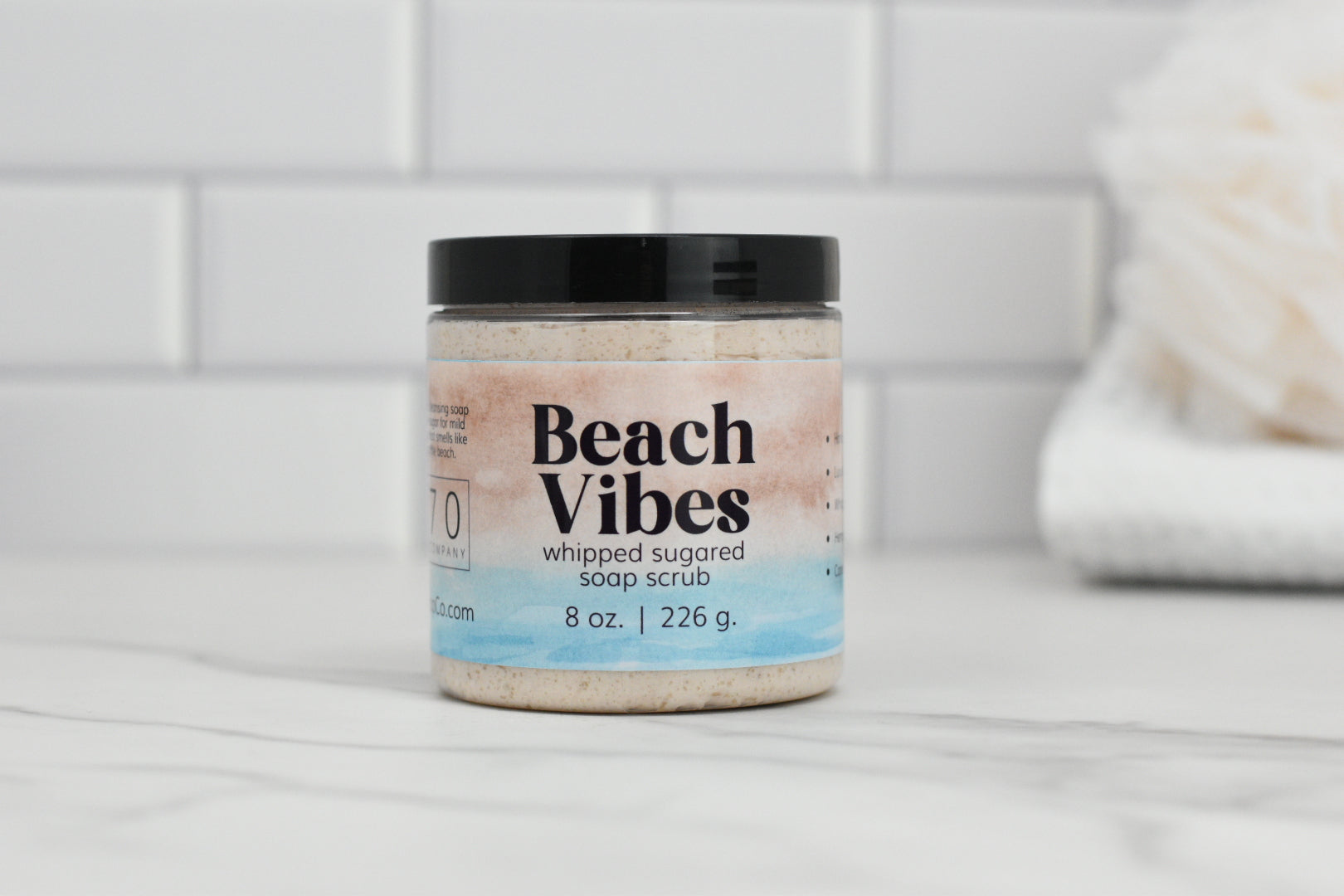 A jar of 570 Soap Co.'s Beach Vibes Sugared Whipped Soap Scrub, featuring blue and black text on the label, rests elegantly on a marble countertop against a white subway tile backdrop. In the blurred background, a white loofah and towel complete the scene, inviting you to a tropical escape into exfoliation elegance.