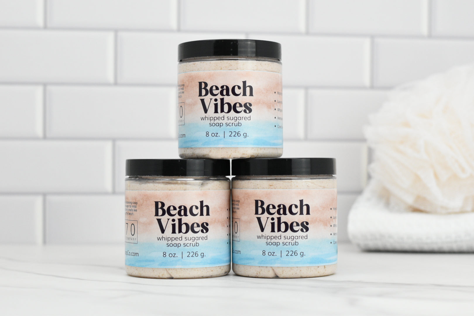 Three jars of Beach Vibes Sugared Whipped Soap Scrub from 570 Soap Co., embodying Exfoliation Elegance, are stacked on a marble countertop against a white tiled wall. A beige bath pouf graces the background on the right, inviting you to indulge in a serene cleansing ritual.