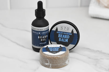 A selection of natural beard grooming products on a marble surface, featuring a dropper bottle of Date Night beard oil with a hint of Kentucky Bourbon fragrance, a customizable scented tin labeled Beard Balm from the Outlaw Collection, and a wrapped bar labeled Beard Wash, all by 570 Soap Co.