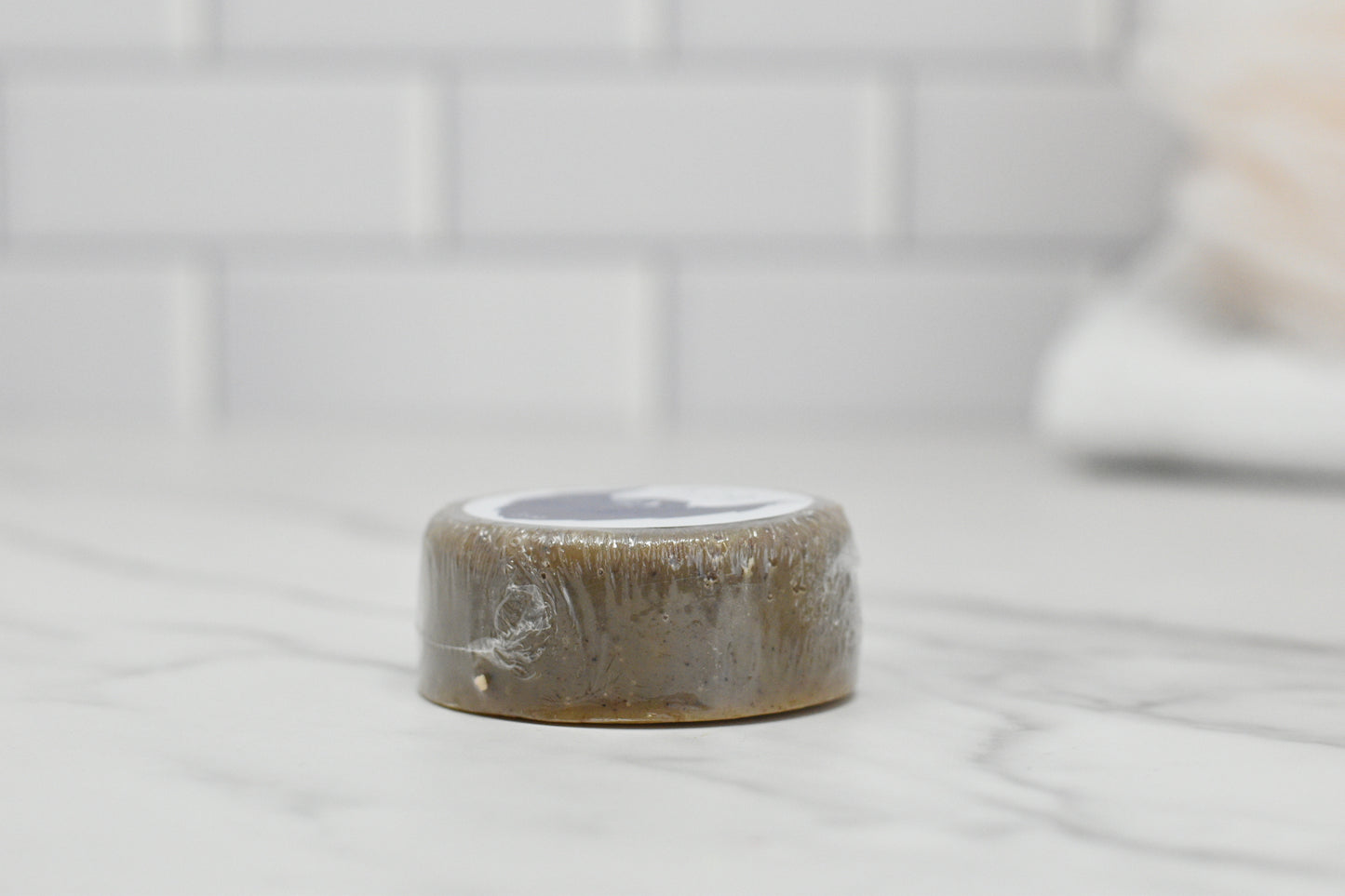 A compact, round bar of Beard Wash - Beard Shampoo enriched with saponified coconut oil and wrapped in plastic is positioned on a marble countertop. The clean and minimalistic setting is accentuated by a white tiled wall in the background, highlighting the 570 Soap Co. brand's attention to detail.