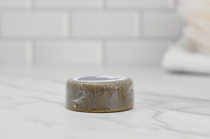 A compact, round bar of Beard Wash - Beard Shampoo enriched with saponified coconut oil and wrapped in plastic is positioned on a marble countertop. The clean and minimalistic setting is accentuated by a white tiled wall in the background, highlighting the 570 Soap Co. brand's attention to detail.