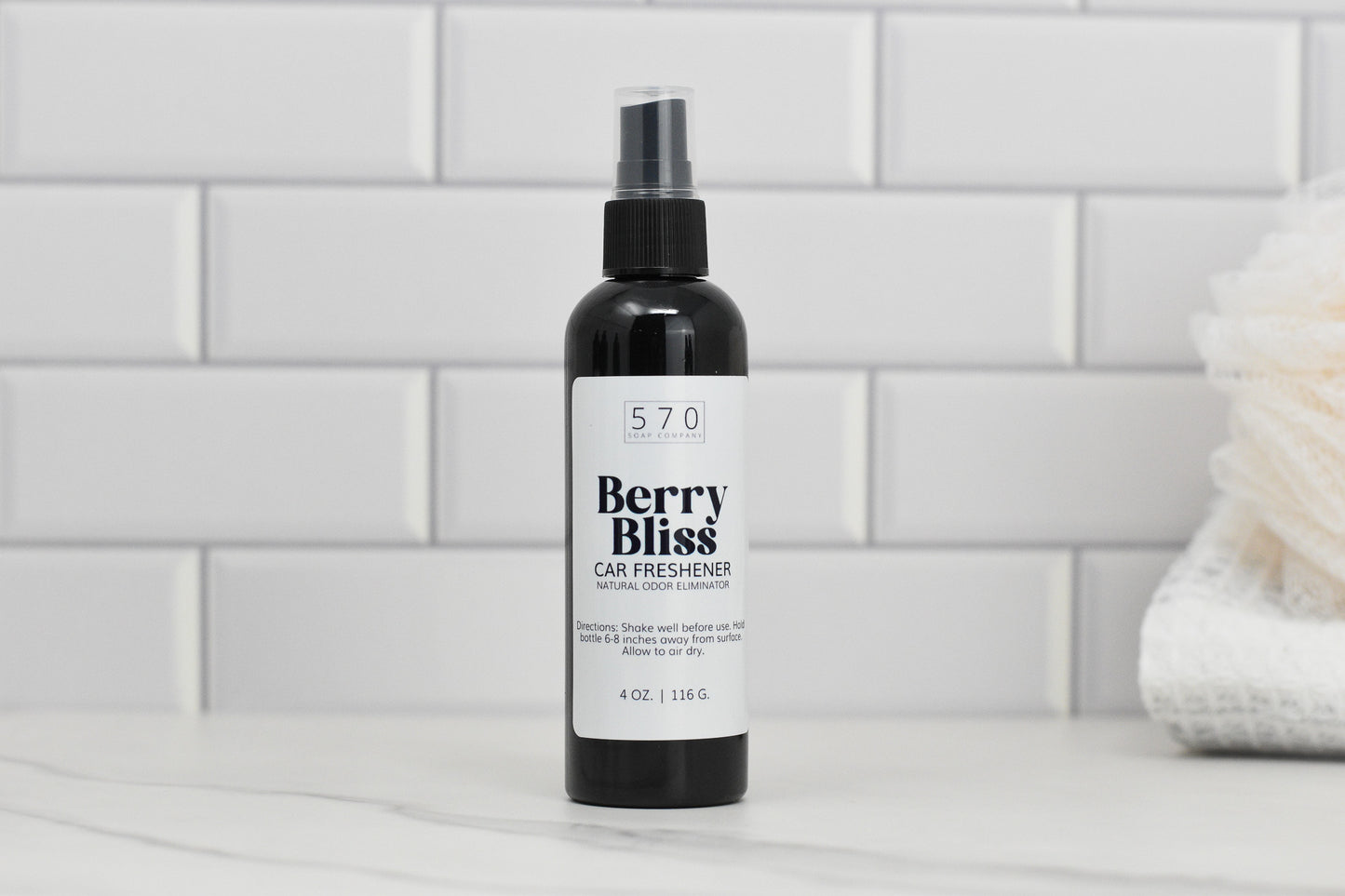 A black spray bottle labeled Linen & Room Spray from 570 Soap Co., a natural odor eliminator, is perched on a marble countertop. The background features white subway tile and a folded white towel, adding a touch of elegance to this delightful fragrance.