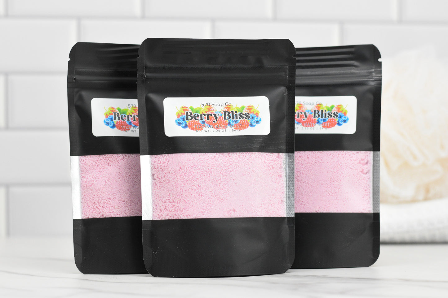 Set against a white tiled background, three black resealable pouches from 570 Soap Co. are labeled as Berry Bliss Fizzing Bath Dust, promising a luxurious experience. Each pouch contains pink bath powder infused with skin-softening ingredients for the ultimate pampering treat.