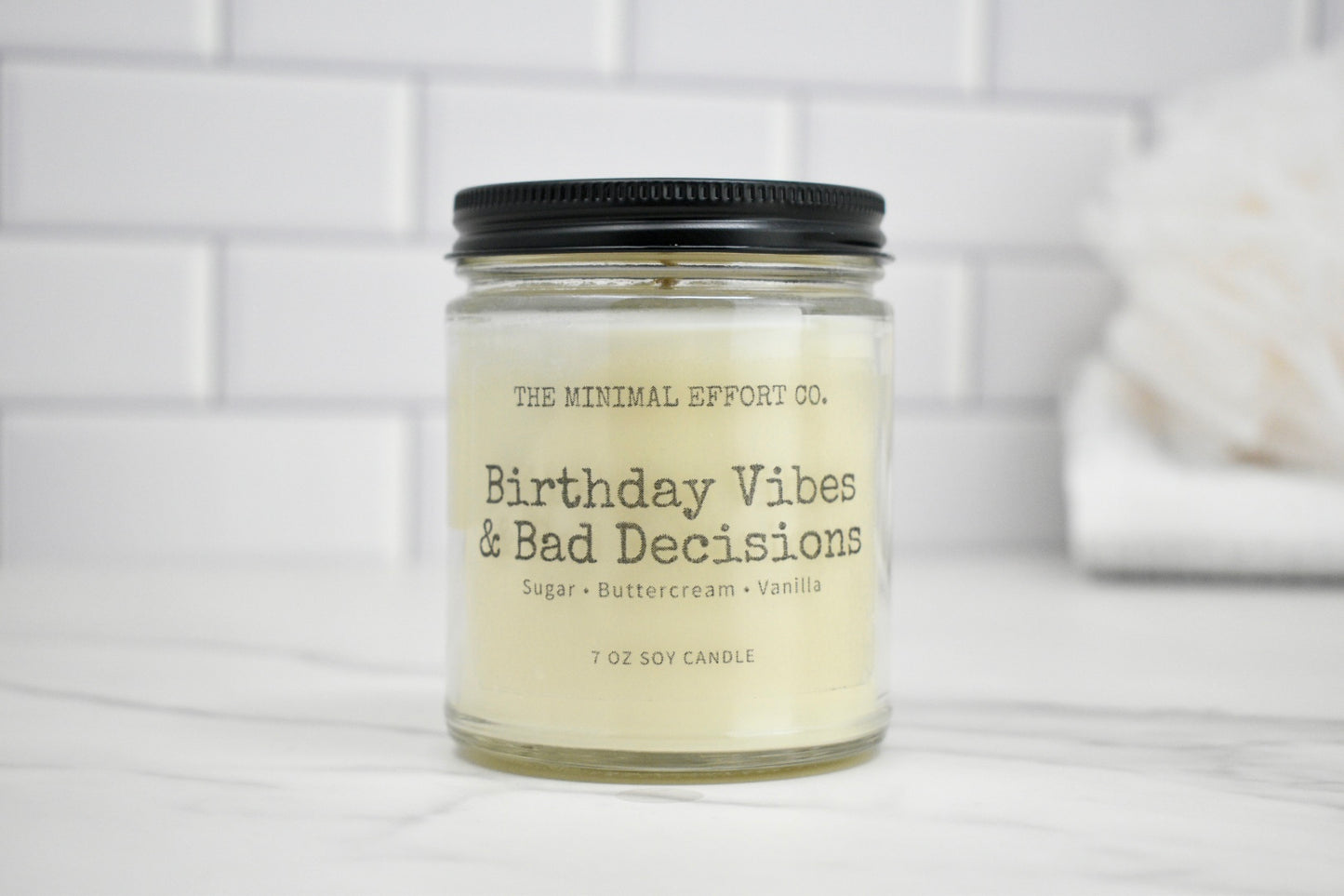 Candle jar labeled 'Birthday Vibes & Bad Decisions' by The Minimal Effort Co. on a white surface with a tiled wall background.