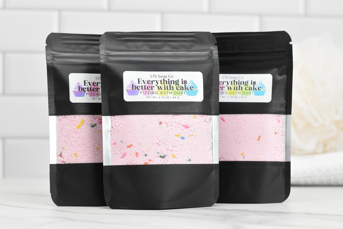 Three black stand-up pouches labeled with the 570 Soap Co. logo and the product name, Birthday Cake Fizzing Bath Dust, display pink fizzy bath dust infused with birthday cake fragrance oil and colorful sprinkles. They rest on a white tiled surface, with a loofah in the background enhancing the charm of this delightful product.