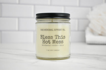 Candle jar labeled 'Bless This Hot Mess' by The Minimal Effort Co. on a white surface with a tiled wall background.